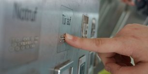 Finger, der auf Aufzugknopf drückt: "Tür auf" ist mit Braillezeichen beschriftet
