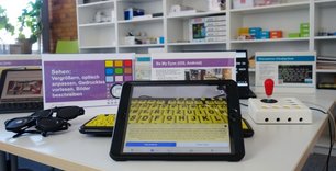 iPad mit der Bildbeschreibungs-App Be My Eyes, die eine schwarz-gelbe Kontrasttastatur für Menschen mit Sehbehinderung beschreibt. Im Hintergrund andere assistive Technologien