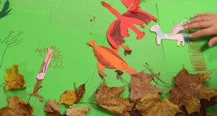 von Kindern gebastelte Kulisse für einen Stop-Motion-Film: Buntes Papier und Laub als Hintgrund, eine Kinderhand schiebt einen aus Papier ausgeschnittenen Drachen  ein Stück für die nächste Aufnahme weiter.