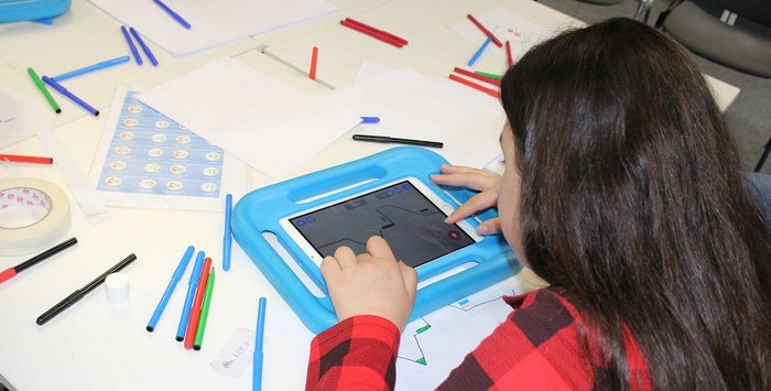 Mädchen mit Tablet und der App Draw Your Game. Vor ihr auf dem Tisch weiße Blätter und bunte Stifte, mit denen sie die Level zeichnet
