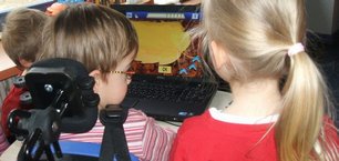 Ein Mädchen und ein Junge vor einem Lernspiel am Computer. Der Junge sitzt im E-Rollstuhl.