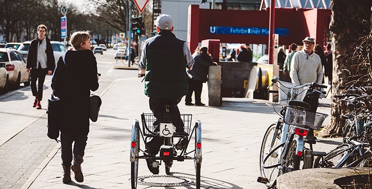Straßenszene: Mann fährt auf Dreirad, neben ihm geht eine Frau